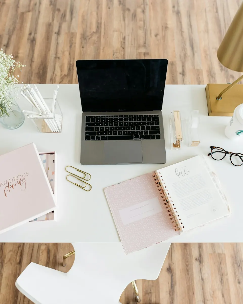 Overhead view of a laptop, notebooks and glasses on a white desk.
